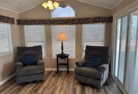 Two gray recliners with blue pillows face a small round table with a lamp, set in front of three windows with blinds in a bright, wood-floored room.
