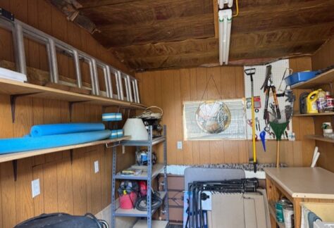 Organized shed with wooden walls, shelves holding various tools and supplies, ladders, folding chairs, and a pegboard with hanging garden tools and equipment.