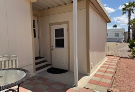A beige manufactured home with a small covered entryway, two doors, a concrete path, and patio furniture beside gravel landscaping and palm trees.