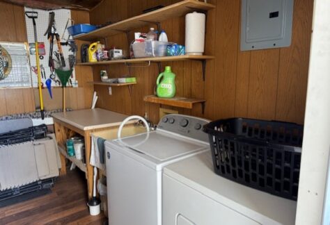 A laundry room with a washer, dryer, black laundry basket, shelves holding cleaning supplies, and tools hanging on a pegboard.