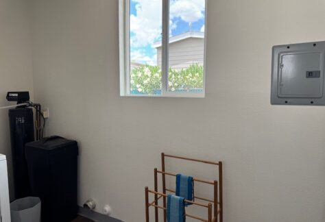 A small laundry room with a window, electrical panel, black water softener unit, trash bin, and a wooden rack holding two blue towels.