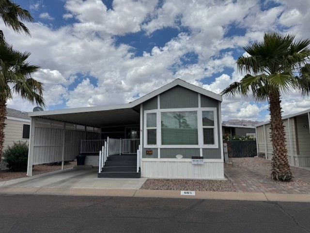 Single-story manufactured home with covered carport, front steps, gray and white exterior, palm trees, and cloudy sky in the background.