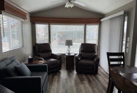 A living room with two brown recliners, a dark sofa, a side table with a lamp, wood flooring, and large windows with blinds letting in natural light.