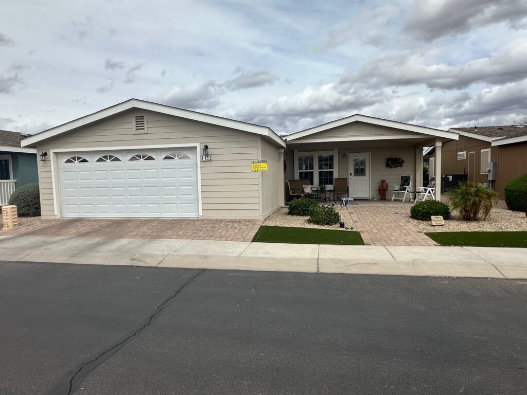 Single-story house with beige siding, white garage door, small front porch, and paved driveway. Lawn with shrubs and "For Lease" sign. Cloudy sky above.