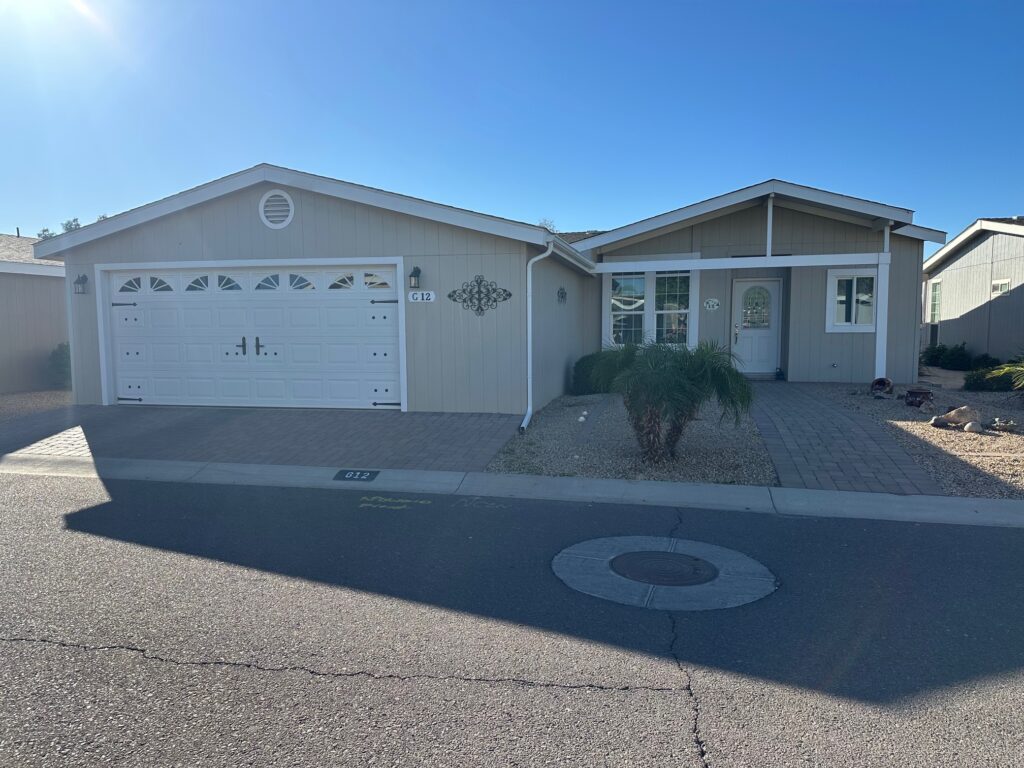 Single-story beige house with a white double garage, small front porch, gravel landscaping, and a palm plant, photographed on a sunny day from the street.