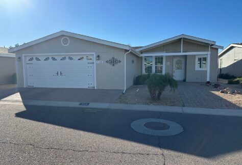 Single-story house with a double garage, beige siding, white trim, small front yard with rocks and a palm plant, viewed from the street on a sunny day.