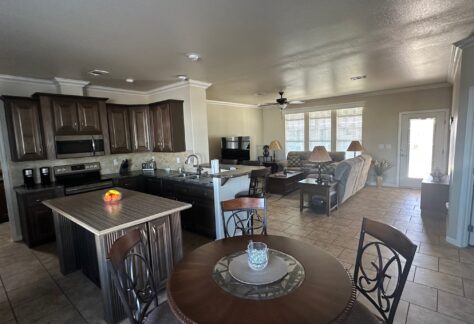 Open-concept kitchen and living room with dark cabinetry, round dining table, island with stools, tile flooring, and large windows letting in natural light.