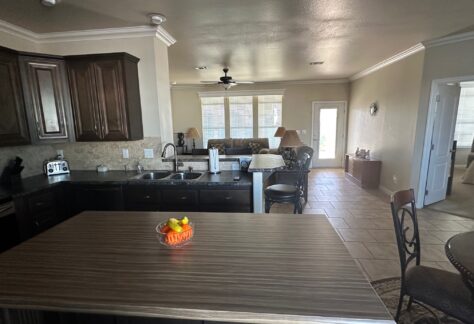 A view of a kitchen with dark cabinets and an island holding a fruit bowl, opening into a living room with a sofa, chairs, and large windows.