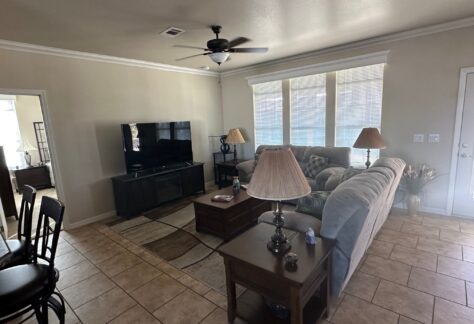 A living room with a sectional sofa, coffee table, lamps, and TV on a stand. Tile flooring and large windows let in natural light. A dining area and another room are visible.