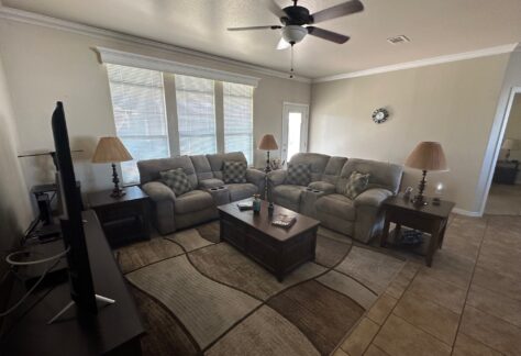 Living room with two beige sofas, a coffee table, two end tables with lamps, a ceiling fan, a wall clock, and a TV on a stand. Large windows let in natural light.