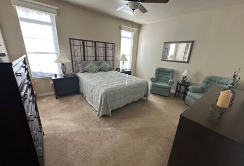 Bedroom with a queen bed, two armchairs, dressers, nightstands, lamps, a ceiling fan, and two windows letting in natural light.