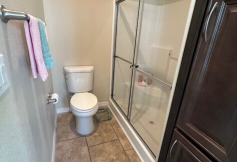 A small bathroom with a toilet, a glass-enclosed shower, tiled floor, towel bar with towels, and a tall dark wood cabinet.