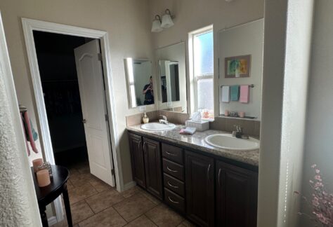 A bathroom with two sinks, dark wood cabinets, a large mirror, wall lights, and various toiletries on the counter. A doorway leads to another room.