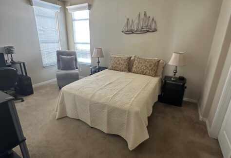 A neatly made bed with beige bedding is centered in a carpeted bedroom, flanked by two nightstands and lamps, with a lounge chair near two large windows.