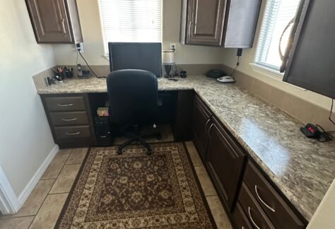 Home office with a U-shaped desk, computer monitor, office chair, brown cabinets, patterned rug, and two windows letting in natural light.