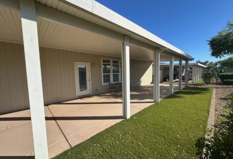 Covered patio with white columns, a concrete floor, a small table and two chairs, adjacent to a house with beige siding and artificial turf along the walkway.