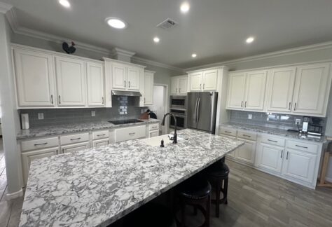 Modern kitchen with white cabinets, stainless steel appliances, gray tile backsplash, and a large marble-patterned island with a sink and four barstools.