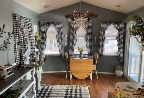 A cozy dining area with a drop-leaf table near three windows, each adorned with black-and-white checkered curtains. A modern chandelier hangs above, and the room has wood flooring.