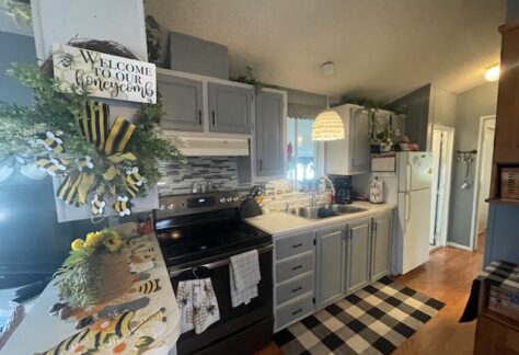 A cozy kitchen with gray cabinets, a white refrigerator, and a black stove. Decor includes a "Welcome to our honeycomb" sign and bee-themed accents. A checkered rug lines the wooden floor.