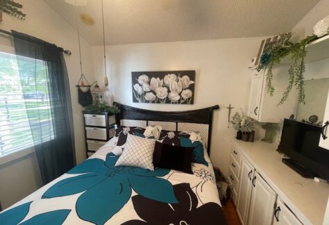 Bedroom with a bed featuring blue and black floral bedding. Wall art of white flowers above the bed. White cabinets and decor on the right. Window with sheer black curtains on the left.