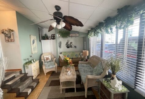 Cozy living room with wicker furniture, floral decor, ceiling fan, and large windows with blinds. Stairs lead to a higher level. Green and beige color scheme.