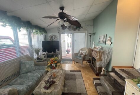 A cozy living room with a wicker sofa, a coffee table, a TV, and a ceiling fan. Green walls are adorned with floral decor. Natural light enters through large windows with blinds.