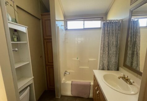 Small bathroom with a white sink, gold faucet, bathtub with shower curtain, wooden cabinets, shelving unit, and a window above the tub.