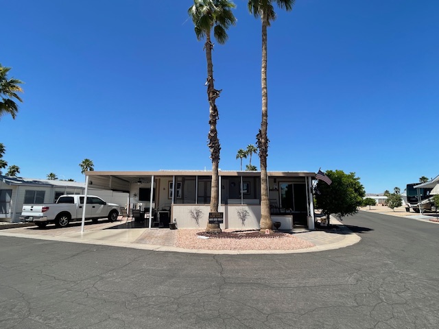 A single-story manufactured home with a carport, two tall palm trees in front, set on a corner lot in a sunny residential area.