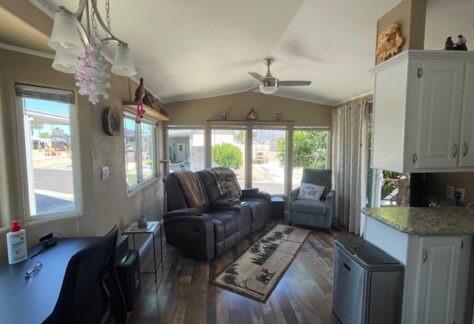 A cozy living room with large windows, a recliner sofa, an armchair, a small desk, and a kitchenette; sunlight streams in, highlighting wood floors and simple decor.