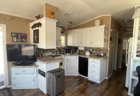 A small kitchen with white cabinets, dark appliances, a wall-mounted TV, and brown walls, featuring wood floors and a countertop with various kitchen items.