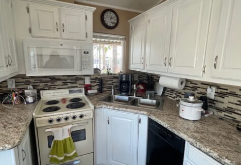 A small kitchen with white cabinets, a white stove, microwave, double sink, dishwasher, coffee maker, and utensil on granite countertops, with a wall clock above the window.