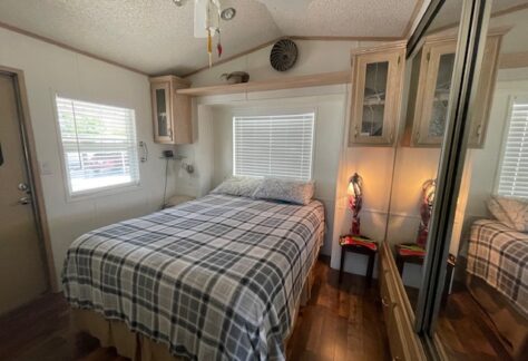 Small bedroom with a plaid-covered bed, wall cabinets, mirrored closet, window blinds, and two decorative lamps on side tables. Wood flooring and ceiling fan are visible.