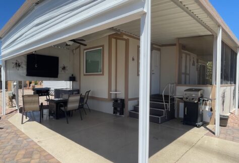 Covered patio area with outdoor dining table, chairs, wall-mounted TV, ceiling fan, grill, and steps leading to a white door, next to a paved walkway.