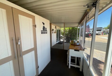 Enclosed porch area with a storage cabinet, small table, and wall sign. Window screens look out onto a yard with an American flag and parked vehicles.
