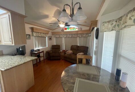 A small living room with two recliners, a ceiling fan, a round dining table with placemats, and a kitchen counter on the left. Blinds cover the windows and doors.