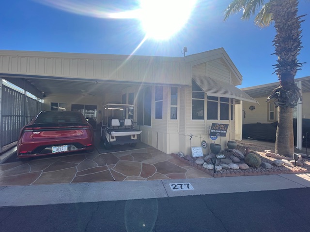 A beige manufactured home with a carport, red car, and golf cart parked outside. Sunlight shines overhead. The address number 277 is visible on the curb. Desert landscaping in front.