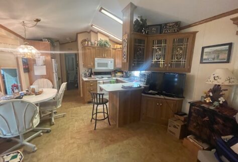 A cozy kitchen and dining area with wooden cabinets, a round dining table, microwave, TV, and various decorations and plants. Natural light enters through a skylight.
