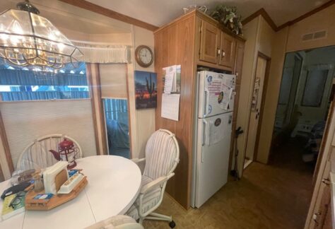 Small kitchen nook with a round white table, four chairs, a hanging light fixture, and a refrigerator next to a hallway in a cozy, wood-accented home.