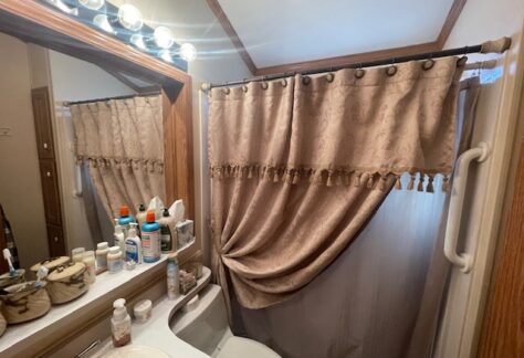A bathroom with a counter holding toiletries, a mirror with lights above, and a shower with beige curtains tied to one side.