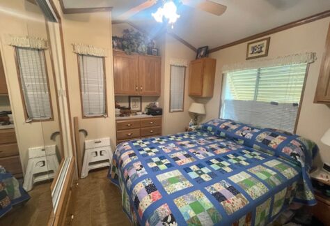 Small bedroom with a colorful patchwork quilt on the bed, wood cabinets above and beside the bed, mirrored closet doors, and two windows with blinds and valances.