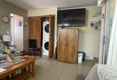 A living room with a TV mounted above a wooden cabinet, a stacked washer and dryer in an open closet, a small desk with a chair, a coffee table, and tiled floors.