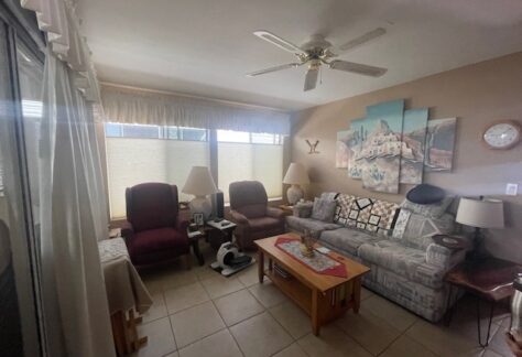 A small living room with a couch, two armchairs, a wooden coffee table, lamps, tile floor, wall art, ceiling fan, and window with blinds letting in natural light.