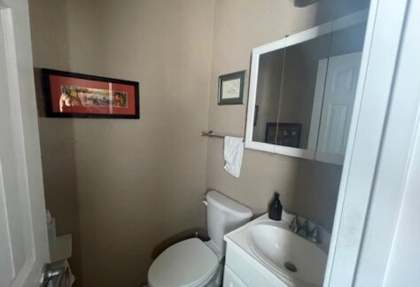 Small bathroom with beige walls, a toilet, white sink vanity, mirrored cabinet, towel rack, framed art, and a dark soap dispenser.