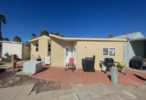 A small, single-story yellow manufactured home with a patio, outdoor chairs, covered grill, and pavers in front, under a clear blue sky.