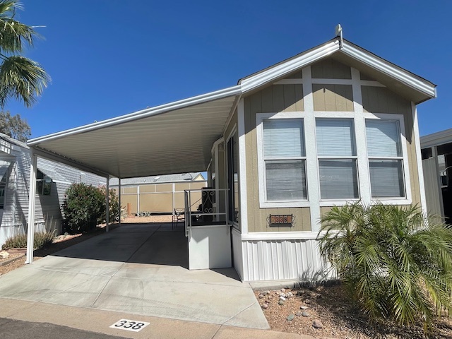 A beige manufactured home with a covered carport, driveway, and small yard with palm plants, under a clear blue sky. The home has several front windows and an address sign.