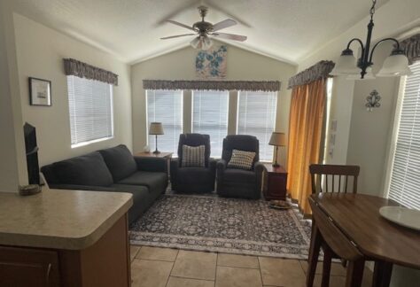 A living room with a sofa, two armchairs, a rug, and two lamps in front of three windows. A ceiling fan and dining table with chairs are also visible.
