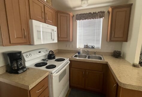 A compact kitchen with wooden cabinets, a white stove and microwave, a double sink under a window with blinds, and a coffee maker on the countertop.