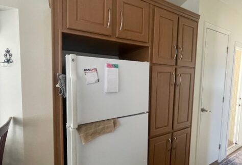 A white refrigerator with papers and a towel attached to it is built into wooden kitchen cabinetry next to a closed door.