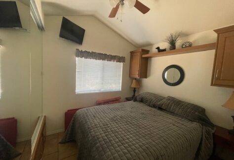 Small bedroom with a neatly made bed, two lamps, a ceiling fan, wall-mounted TV, window with blinds, and mirrored closet doors. Shelves above the bed hold decorative items.
