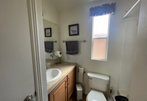 Small bathroom with a sink, countertop, toilet, and shower. A window lets in light, and two towels hang beneath framed pictures above the sink.